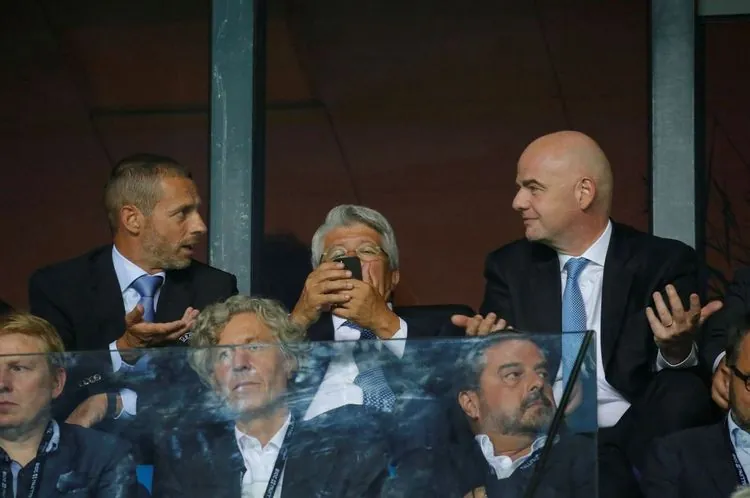 Mehrere Männer in Anzügen sitzen in einer VIP-Loge eines Stadions. Zwei von ihnen, auf der linken und rechten Seite, gestikulieren und sprechen miteinander. Eine Person in der Mitte hält ein Smartphone. Hintergrund unscharf.